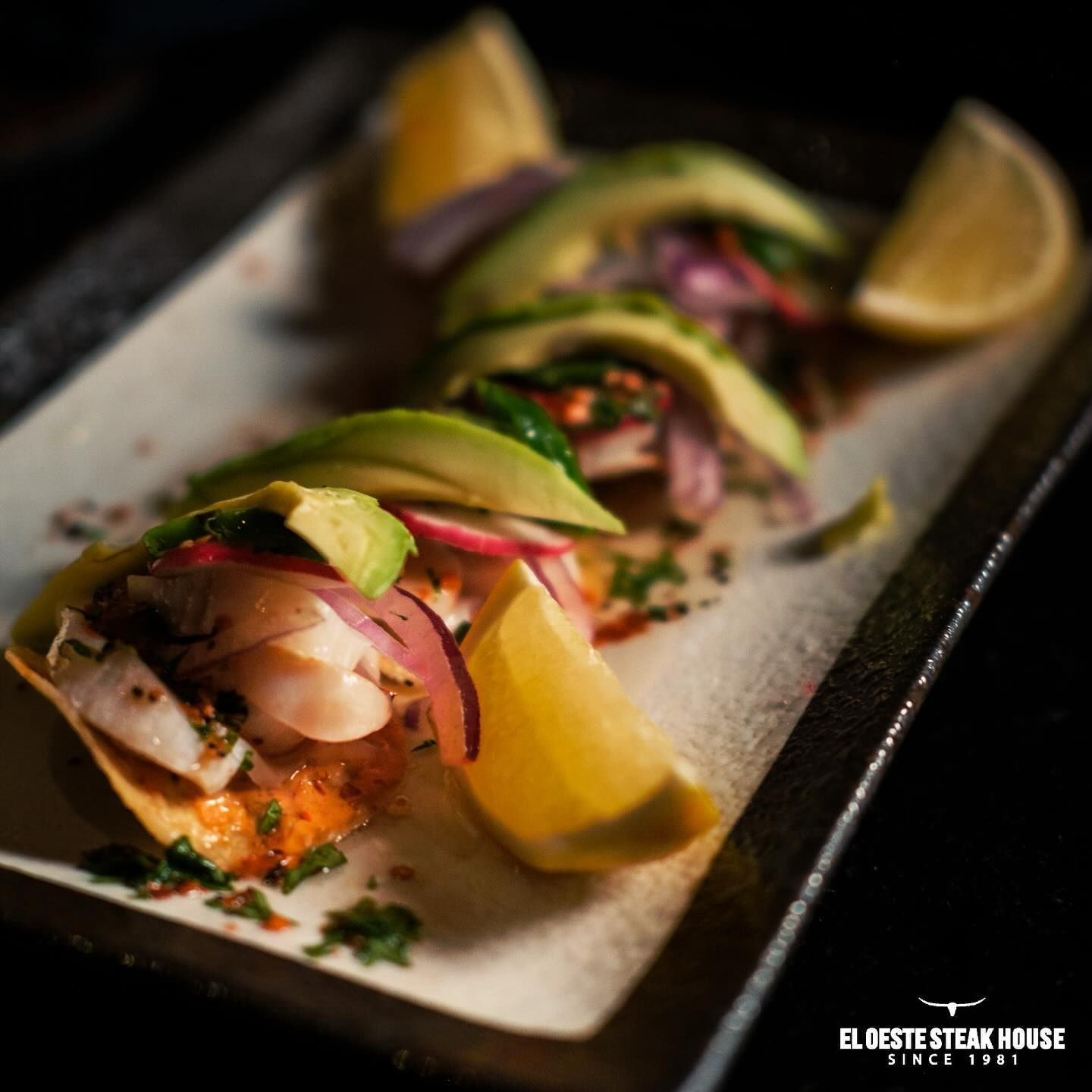 Tostadas cubiertas con ceviche de pescado, aguacate, cebolla roja y gajos de limón en un plato oscuro.