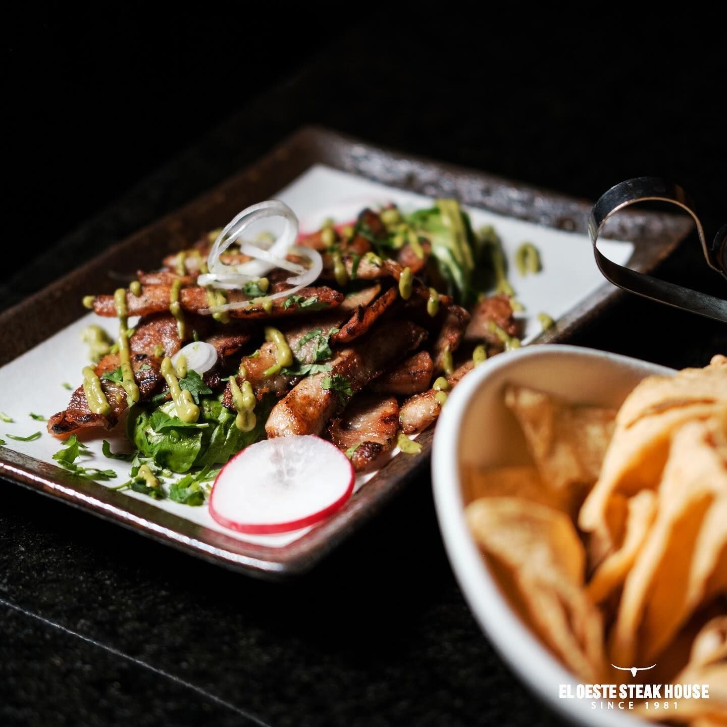 Carne a la parrilla con aguacate, guarnición y totopos en un plato, junto a un tazón de totopos.