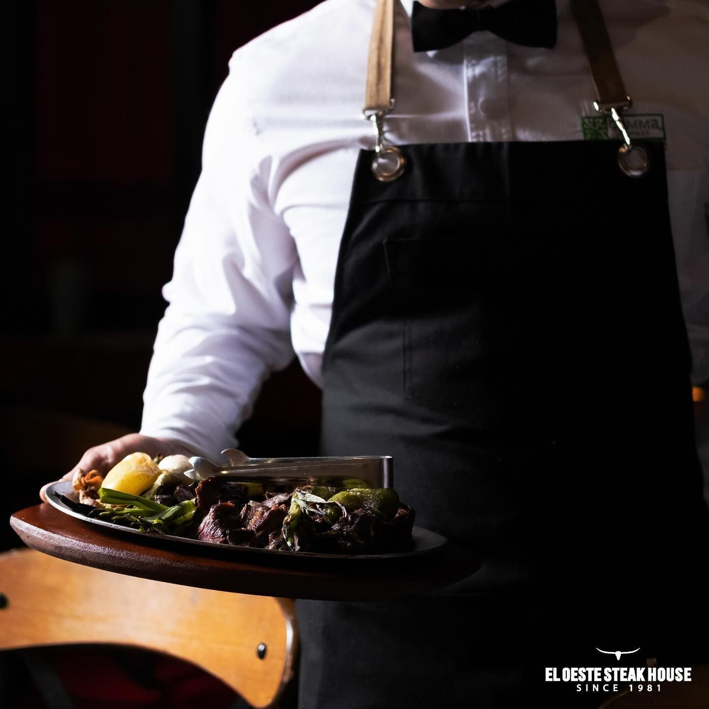 Un camarero sostiene un plato de comida en el restaurante El Oeste Steak House. El camarero lleva un delantal negro.