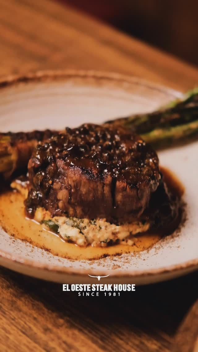 Filete cubierto con salsa de pimienta, servido sobre puré de papas y en un plato, con espárragos.