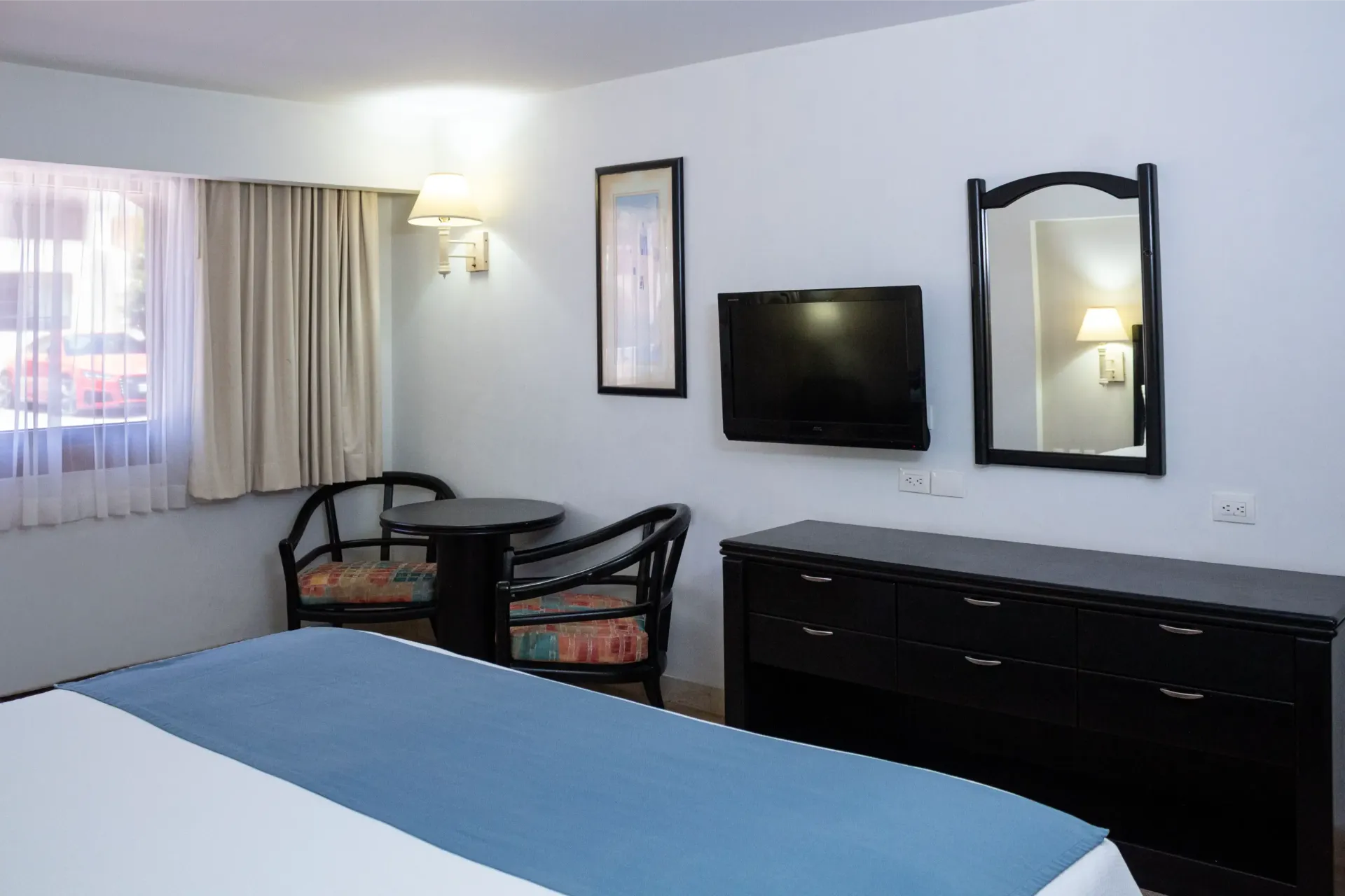 Habitación de hotel con cama, mesa, sillas, cómoda, televisor, espejo y ventana con cortinas.