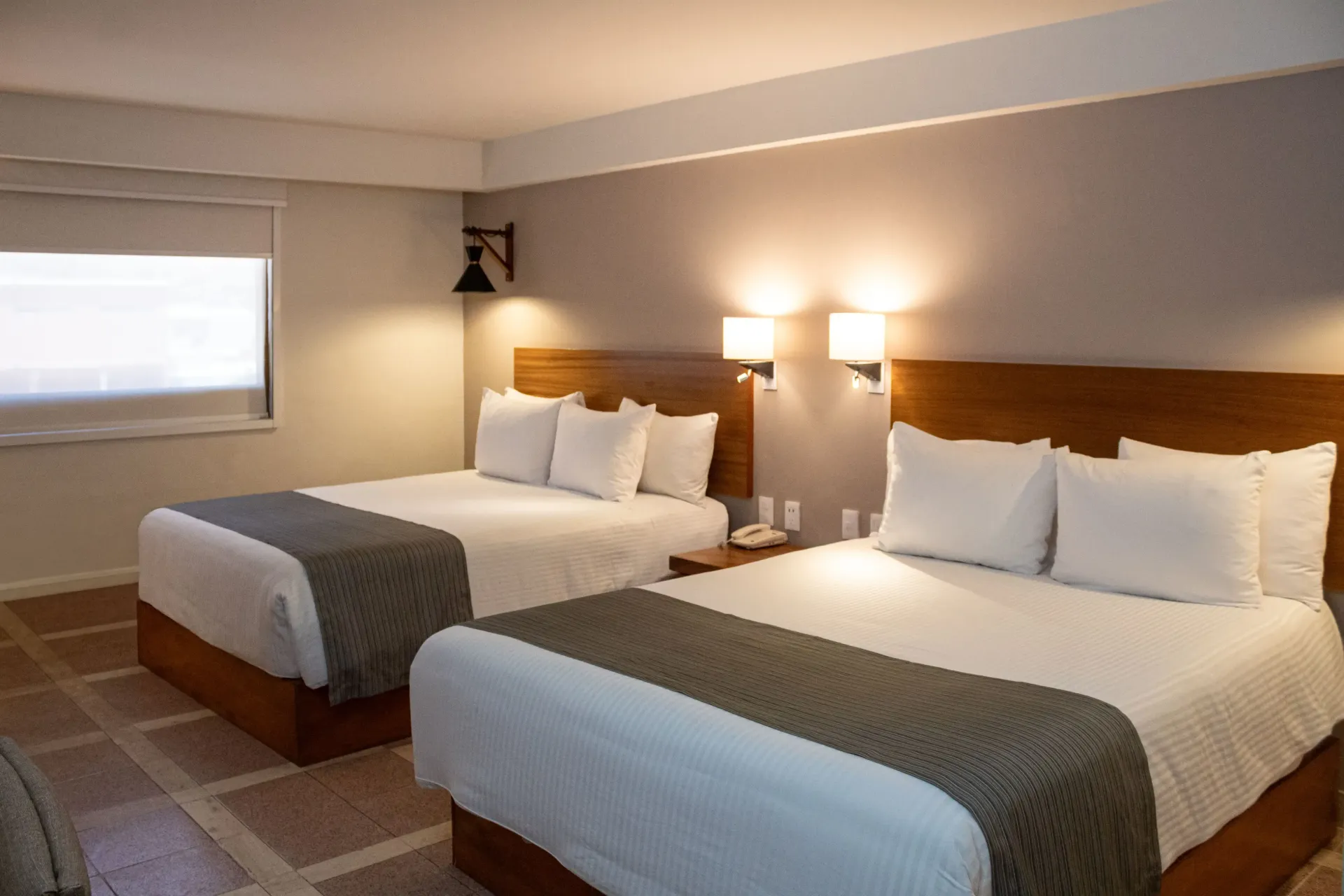 Habitación de hotel con dos camas queen, sábanas blancas, detalles grises, cabeceros de madera y apliques de luz.
