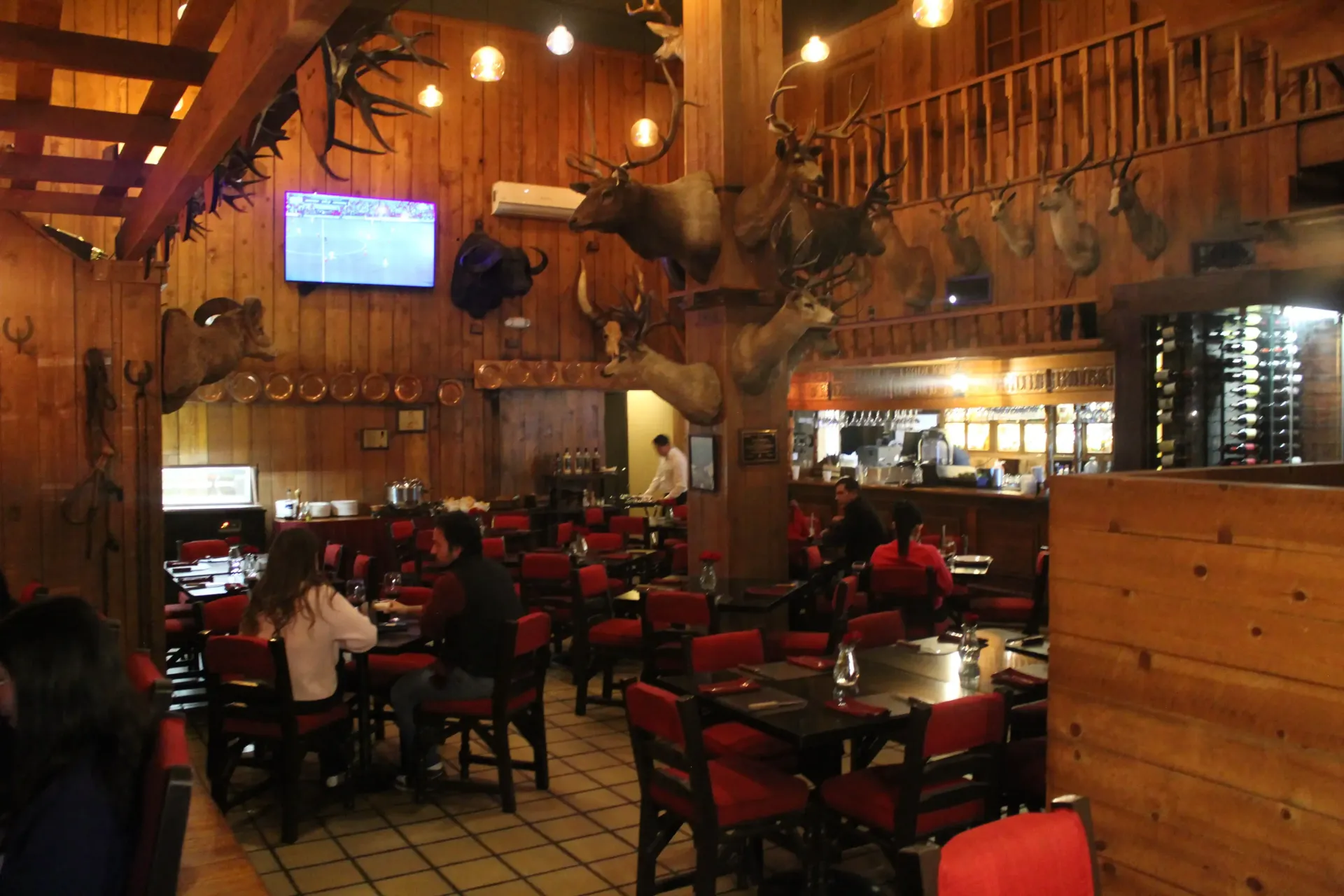 Interior rústico de restaurante con paredes de madera, sillas rojas, animales disecados y clientes.