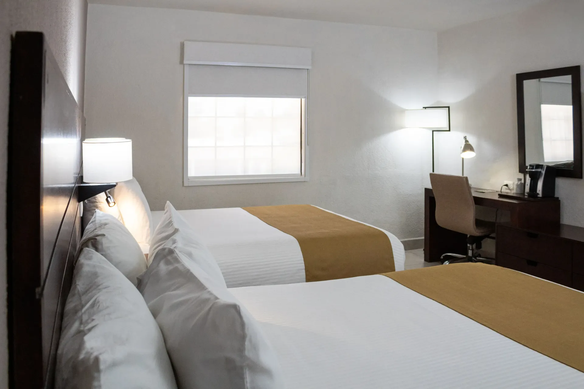 Habitación de hotel con dos camas, un escritorio, una ventana con persianas y un espejo.