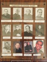 Wall of framed portraits and plaques labeled “Pastors of Notre Dame” in a church or school hallway