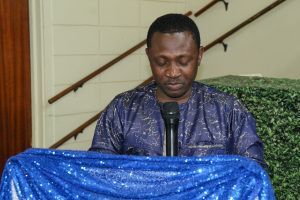 Man speaking at a podium covered with blue cloth, looking down at notes in a room with stairs behind him