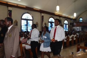 People walking down a church aisle during a service, with pews and arched windows in view.