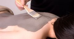 Person's back covered in grey mud as spa technician applies with a brush.