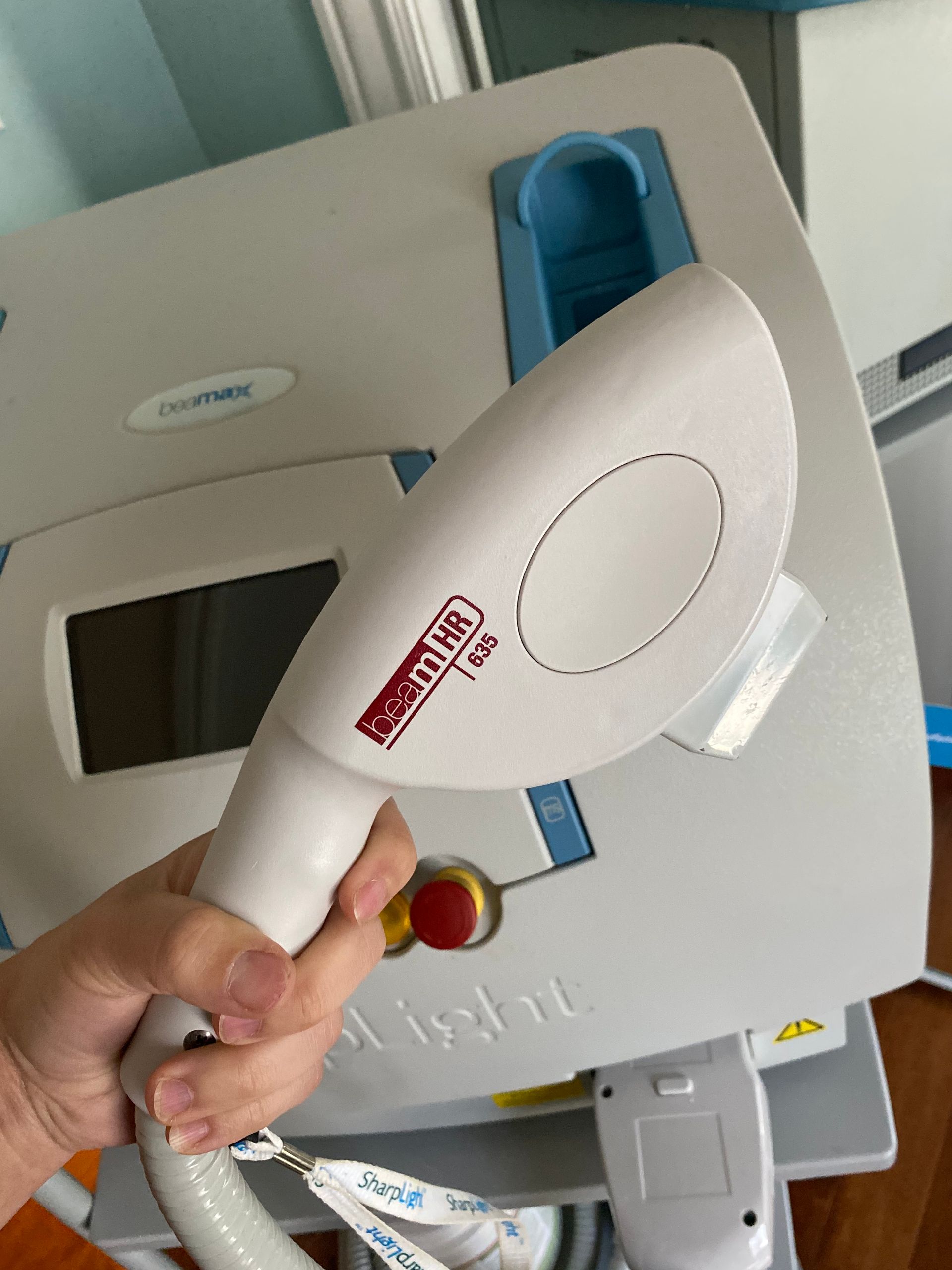 Hand holding a white laser hair removal device in front of a larger machine with a digital display.