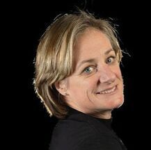 Woman with short blonde hair smiles, looking over her shoulder. Dark background.
