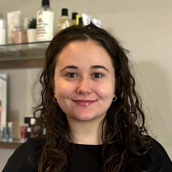 Woman with curly brown hair smiling, standing in front of a shelf of products, in a well-lit space.