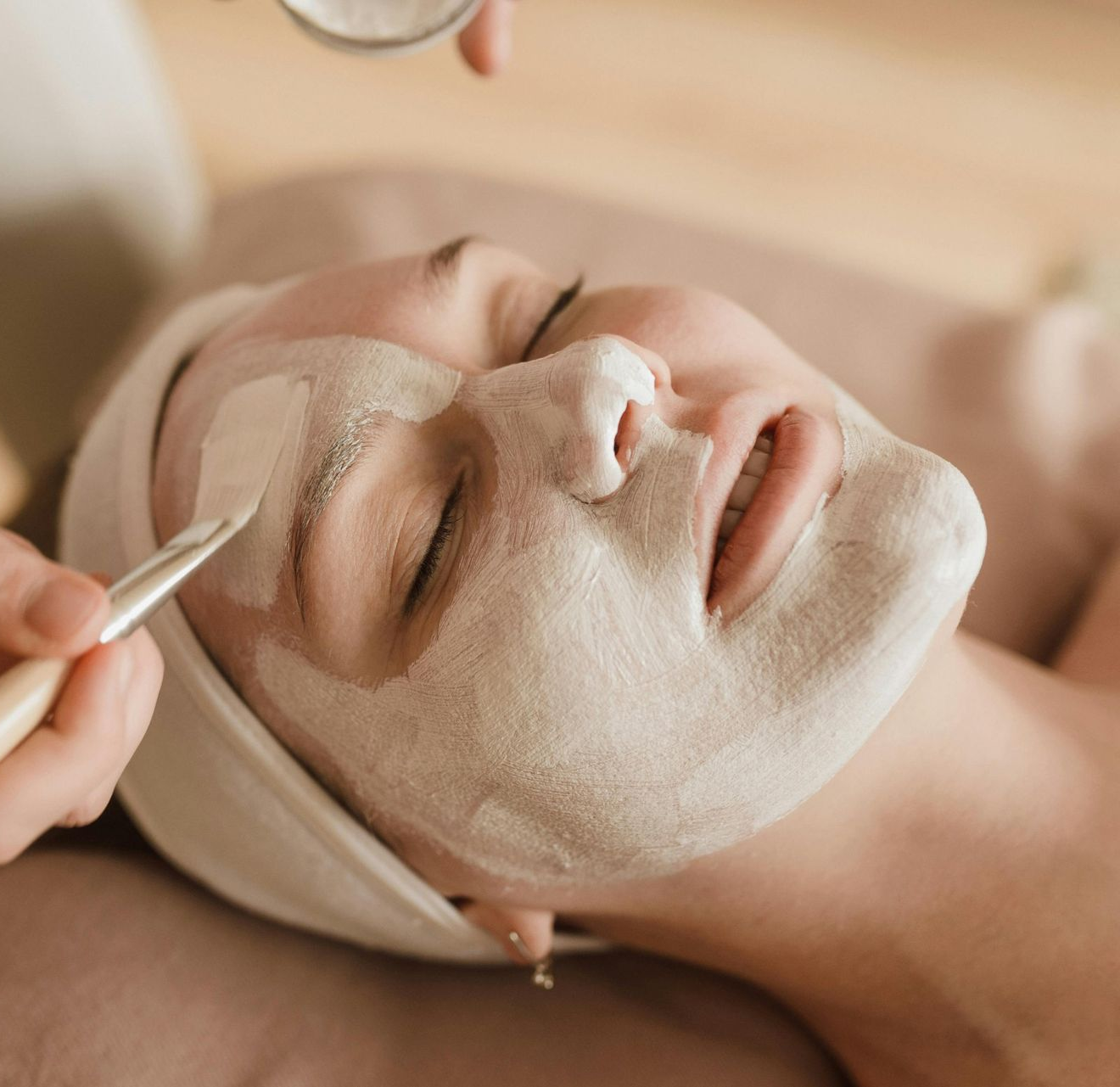 Woman receiving facial mask treatment at a spa, eyes closed, white mask being applied.