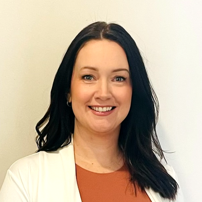 Woman with dark hair smiles, wearing a rust-colored top and white blazer against a white background.