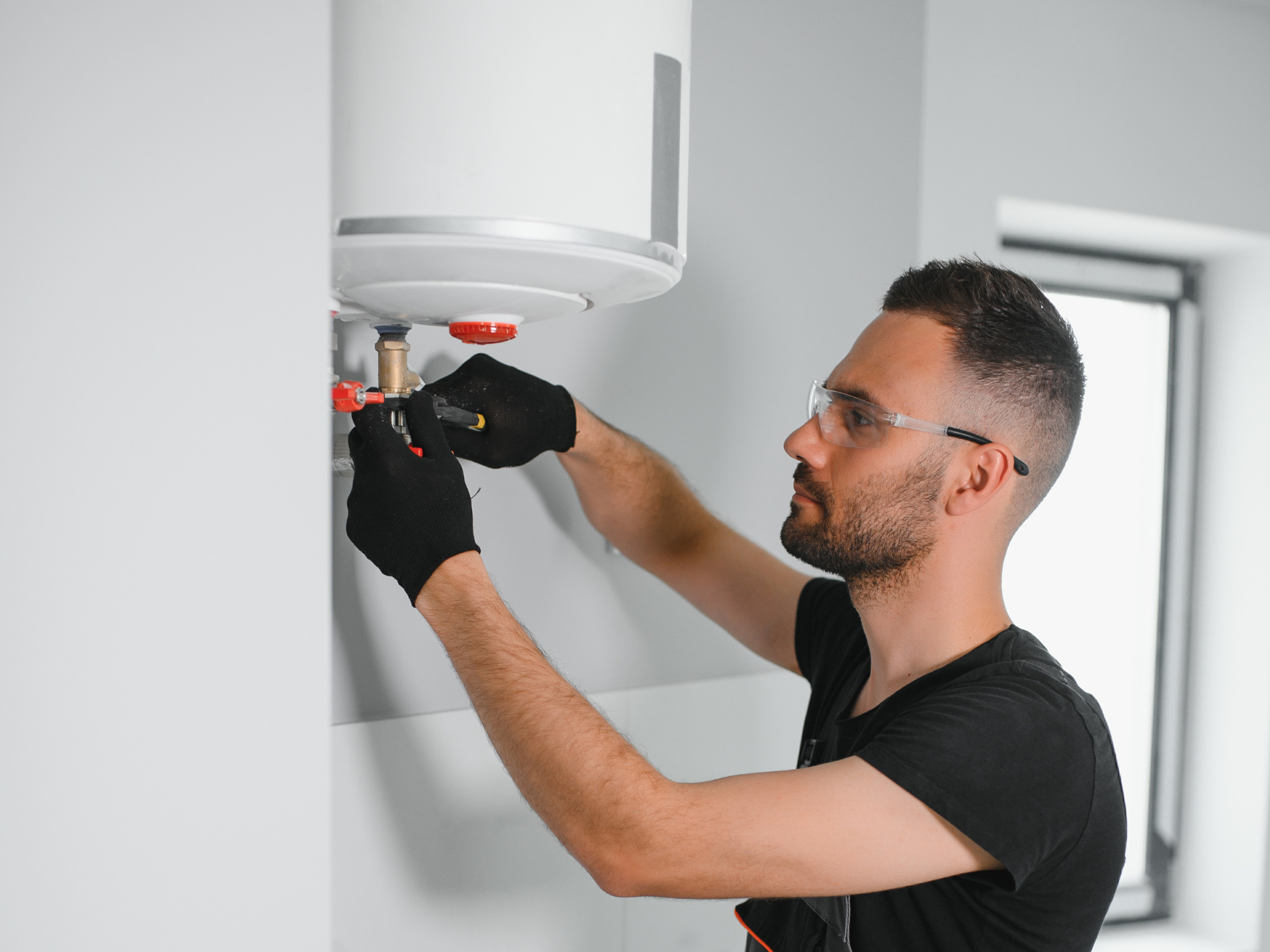 Plumber in black gloves and safety glasses repairing a water heater on a white wall.