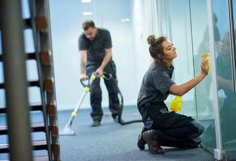 a female cleaning contractor is polishing the glass partition offices whilst In the background a male colleague steam cleans an office carpet in a empty office in between tenants. .The female is smiling