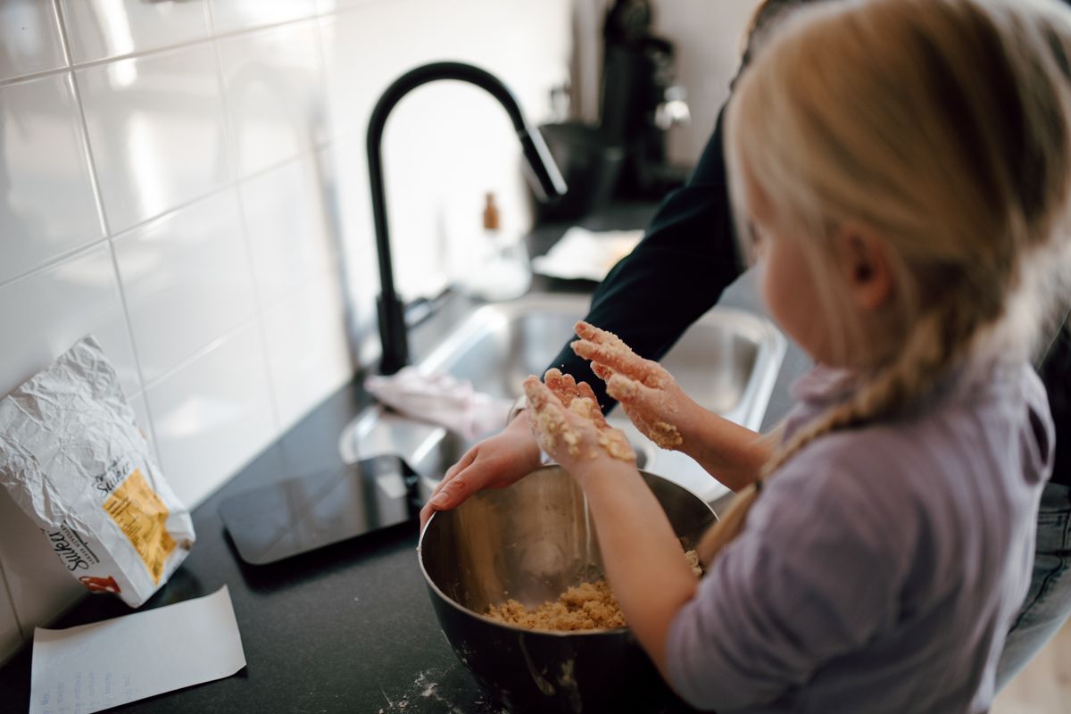 Een volwassene en een kind, beiden met meel bedekte handen, werken samen om deeg te mengen in een metalen kom op het aanrecht.