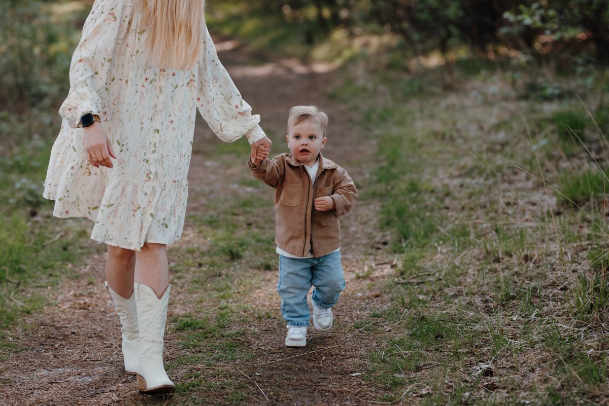 Een vrouw in een lichtgekleurde jurk met bloemenprint en witte laarzen loopt hand in hand met een kind in een bruine jas over een onverhard pad in een bos.