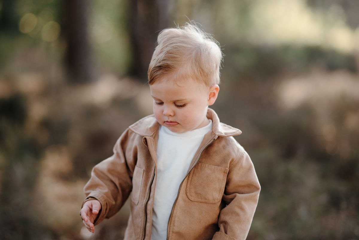 Een jong kind in een beige jasje en een wit shirt kijkt naar beneden terwijl het in een wazig, bosrijk buitenlandschap staat.