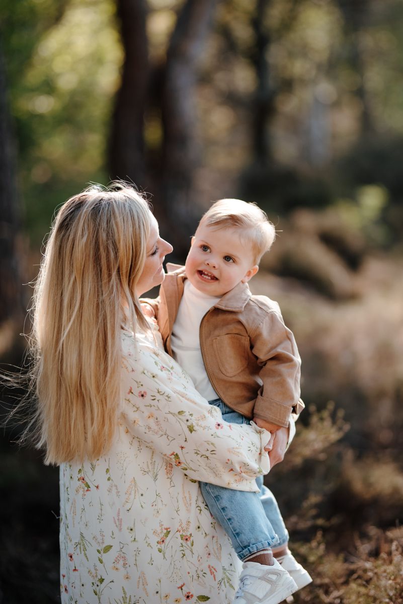 Een blonde vrouw houdt een peuter vast die een beige jasje en spijkerbroek draagt. Ze glimlachen naar elkaar in een zonnig, bosrijk gebied.