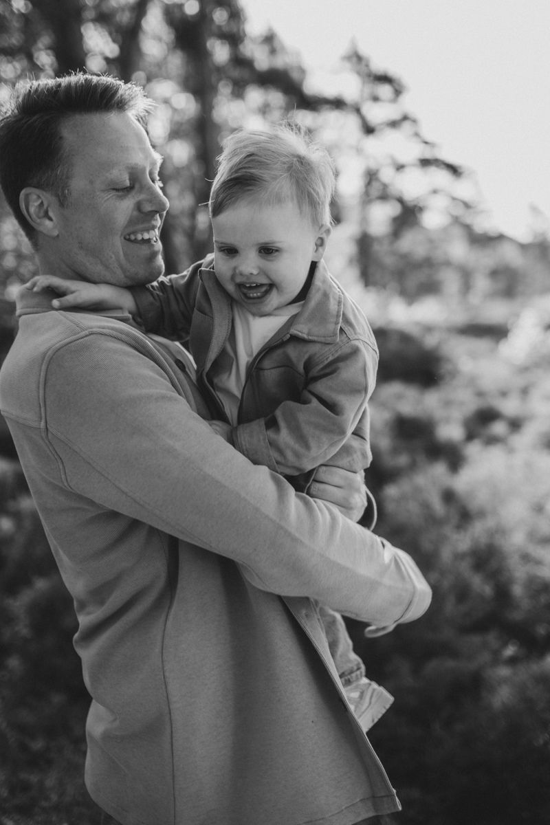 Een persoon lacht terwijl hij een peuter in zijn armen houdt, tegen een wazige, zonovergoten buitenachtergrond.