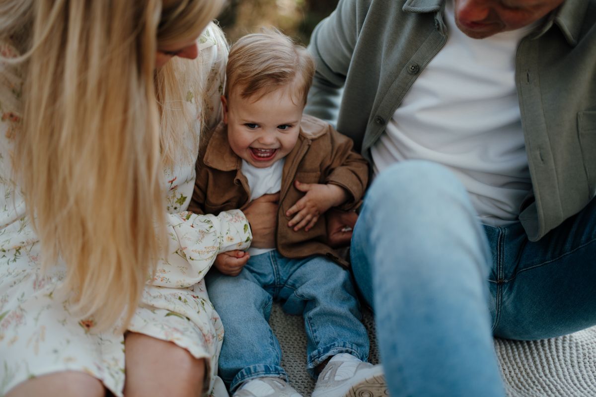 Een ouder in een witte jurk met bloemenprint en een andere ouder in een groen shirt spelen met een lachende peuter in een bruine jas en een blauwe spijkerbroek.