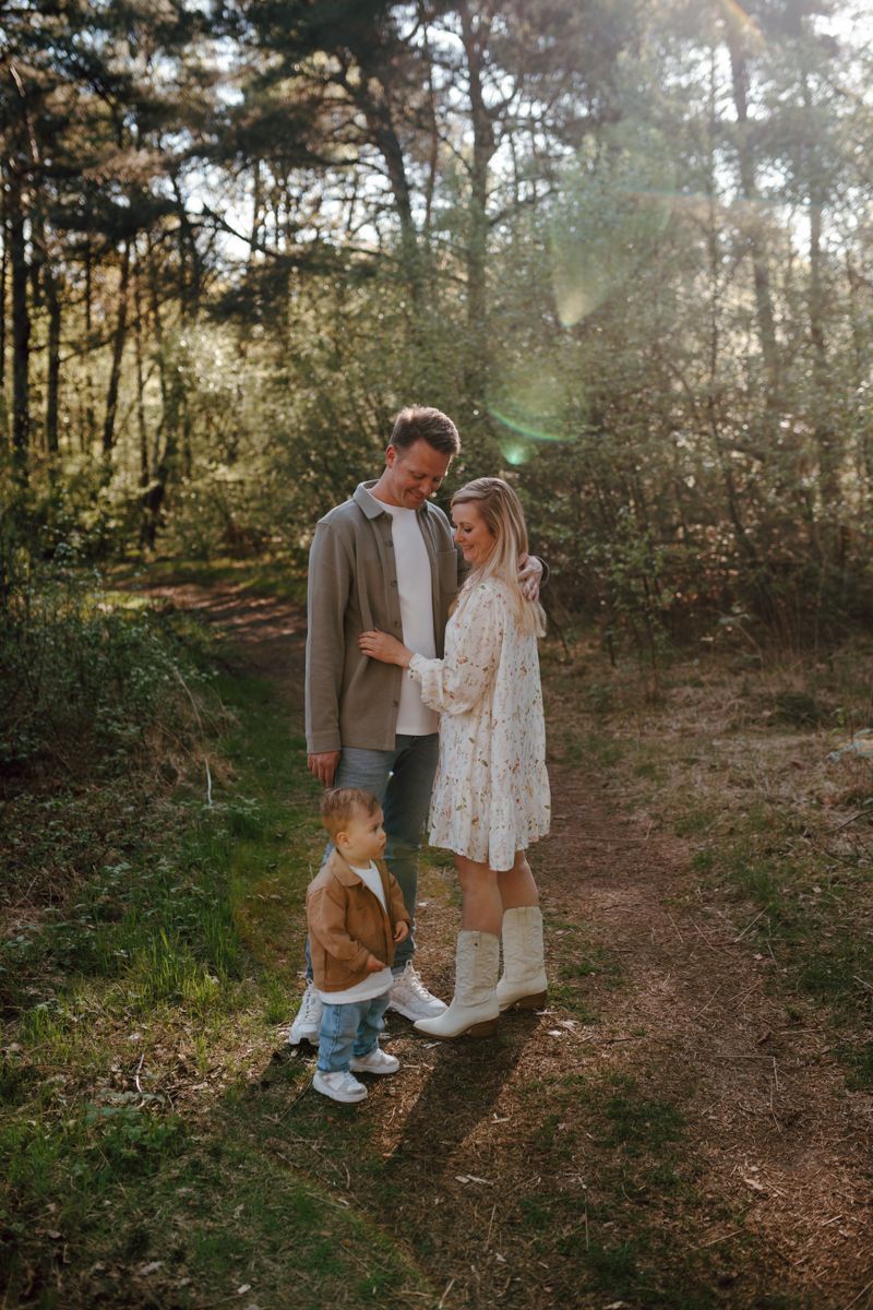 Een gezin van drie staat tijdens het gouden uur op een bospad, waarbij de ouder achterin naar het kind glimlacht.