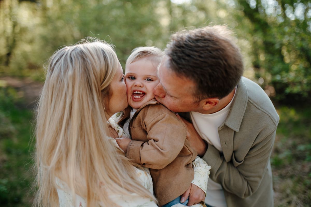 Een jong kind wordt vastgehouden tussen twee volwassenen die het kind kusjes op de wangen geven in een zonnige, bosrijke omgeving.