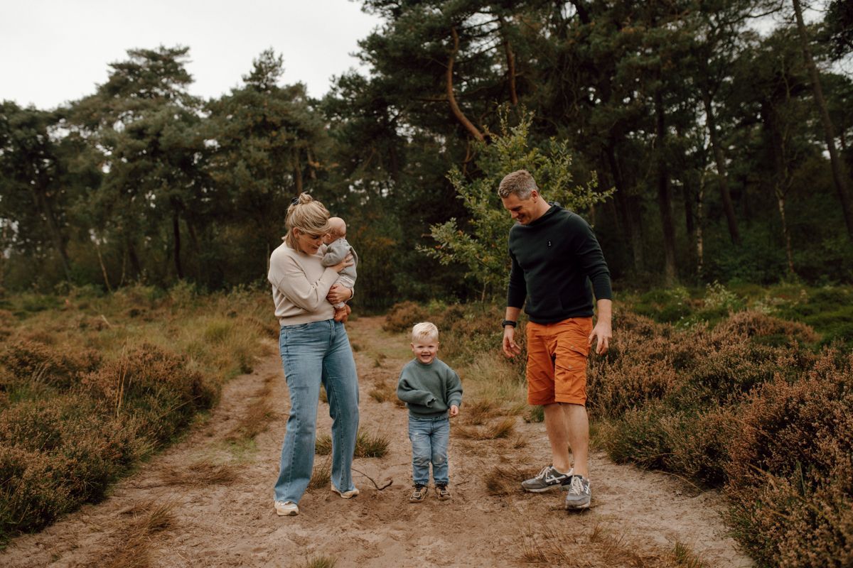 Een gezin van vier loopt over een zandpad in een bos, waarbij een van de ouders een baby vasthoudt en een klein kind voorop loopt.