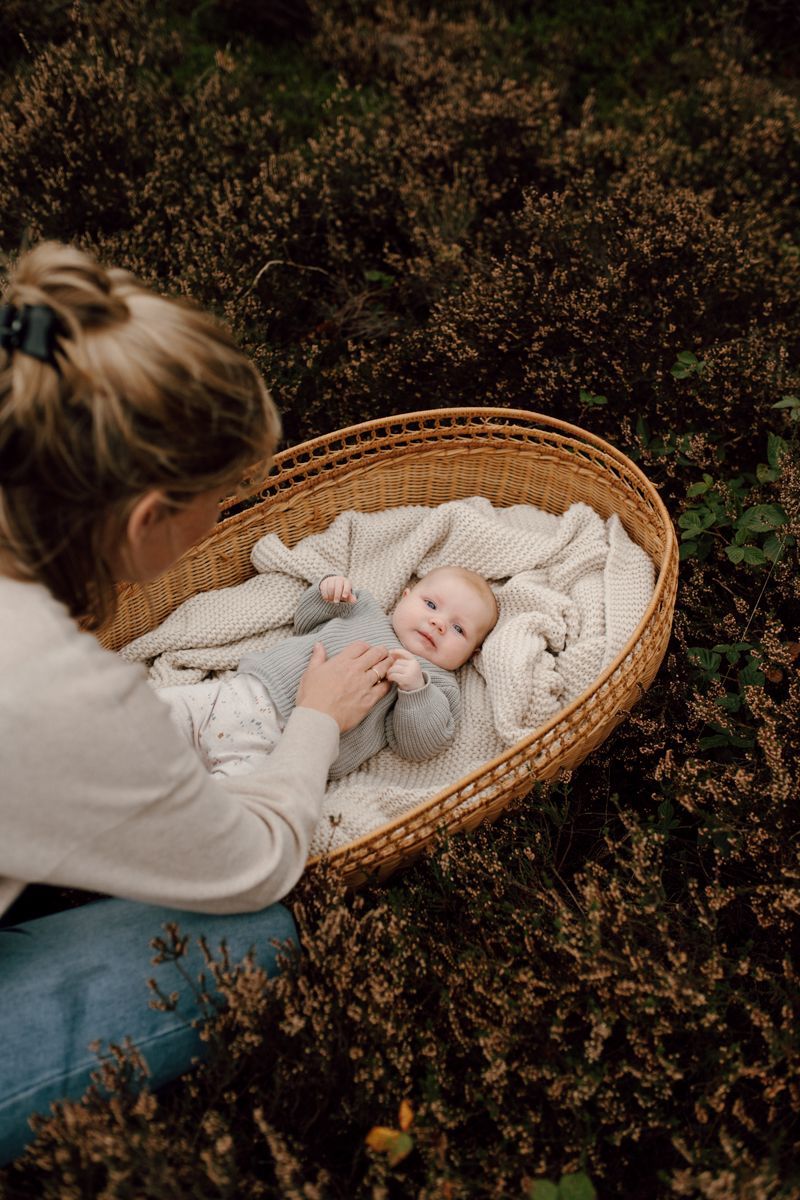 Een persoon met een lichtgekleurd shirt raakt voorzichtig een baby aan die in een rieten mandje ligt, te midden van donker gebladerte.