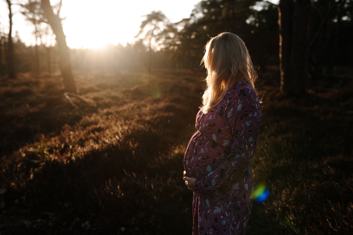Een persoon staat in een zonovergoten bos bij zonsondergang, draagt ​​een jurk met een patroon en houdt haar zwangere buik vast.