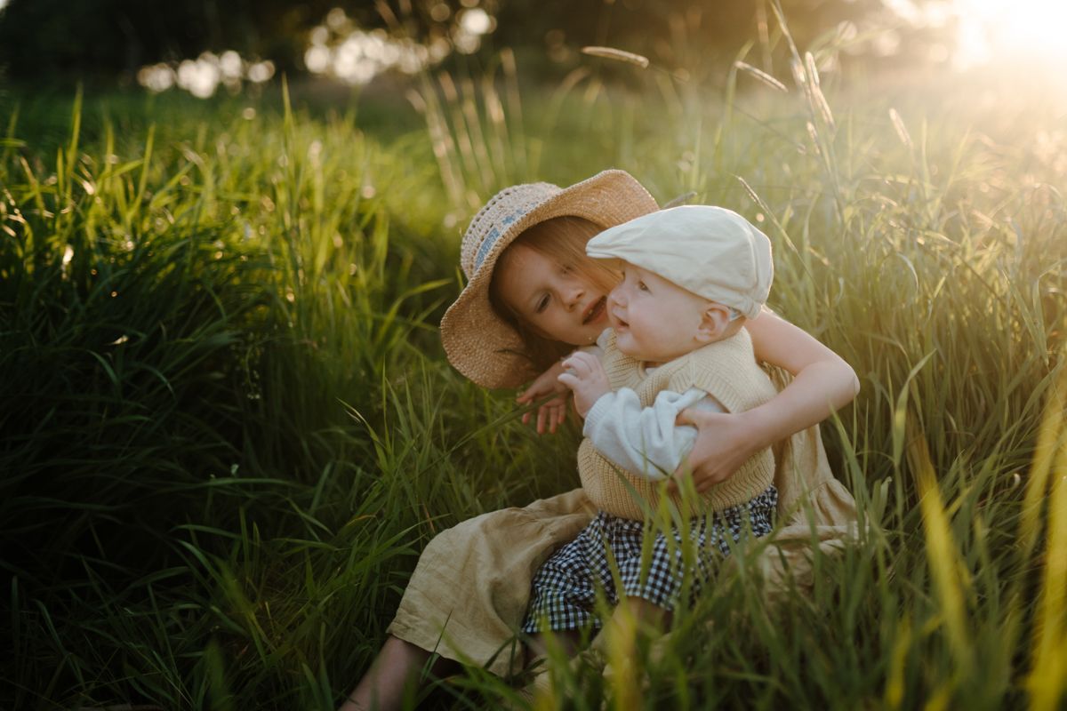 Twee kinderen zitten in het hoge gras in de warme zon, de ene knuffelt de andere die een zonnehoed draagt.