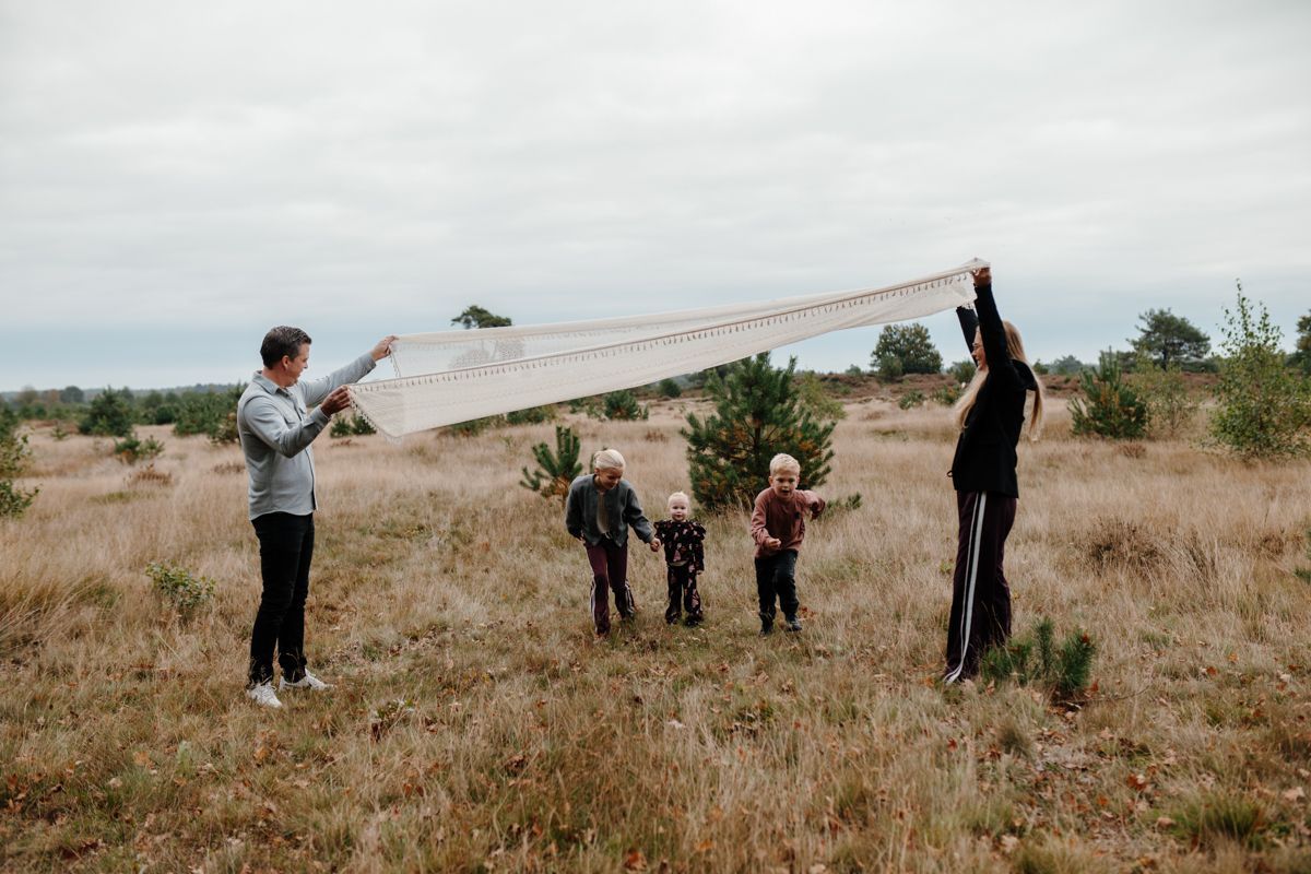 Twee volwassenen houden een lange, bedrukte stoffen banner vast die over een droog, grasveld is gespannen, met drie kinderen die eronder spelen.