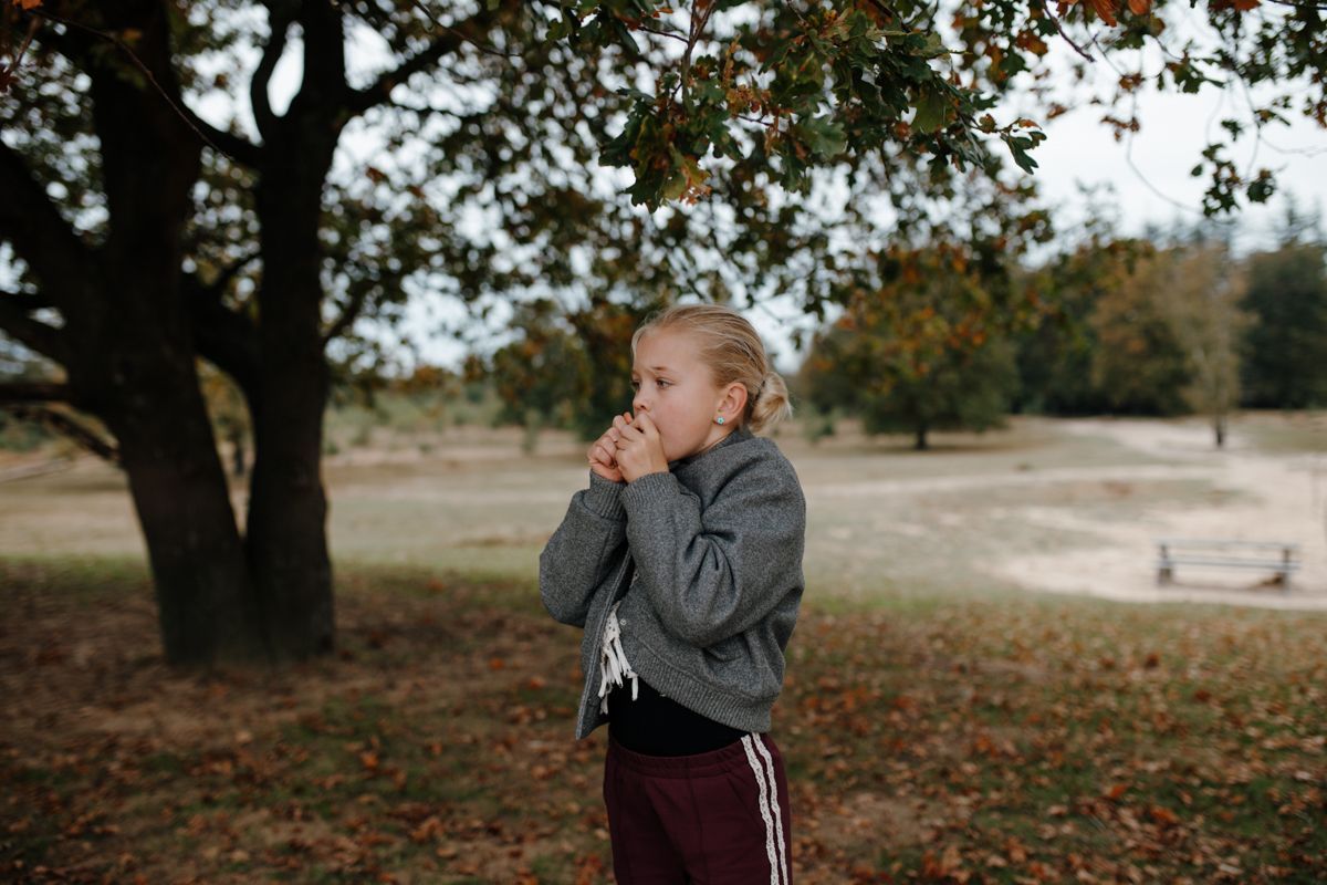 Een kind in een grijze jas staat onder een boom, met de handen bij het gezicht, in een park bedekt met gevallen herfstbladeren tijdens een fotoshoot van het gezin