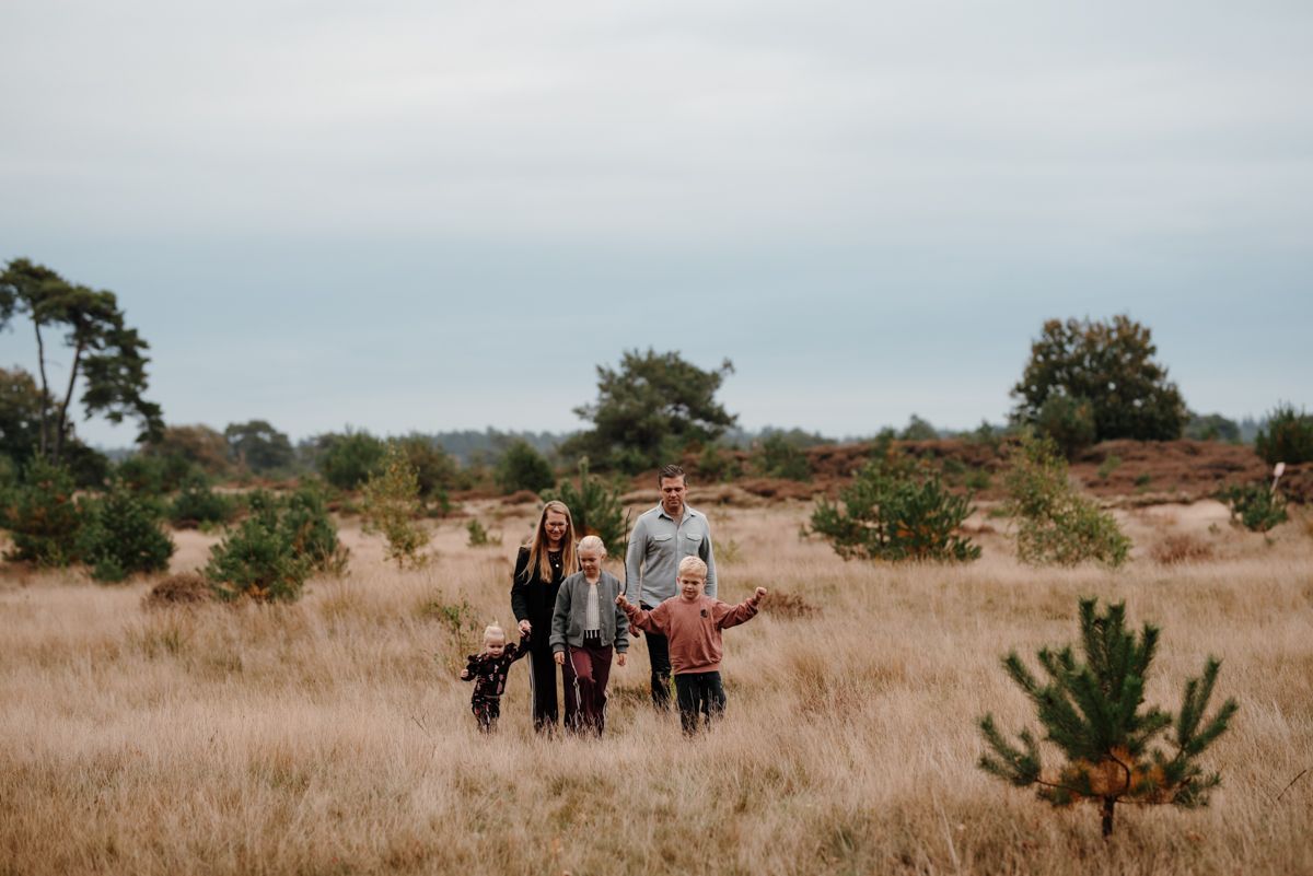 Een gezin van vijf loopt hand in hand door een open, grasrijk landschap onder een zacht bewolkte hemel. Gezinsfotografie in de natuur