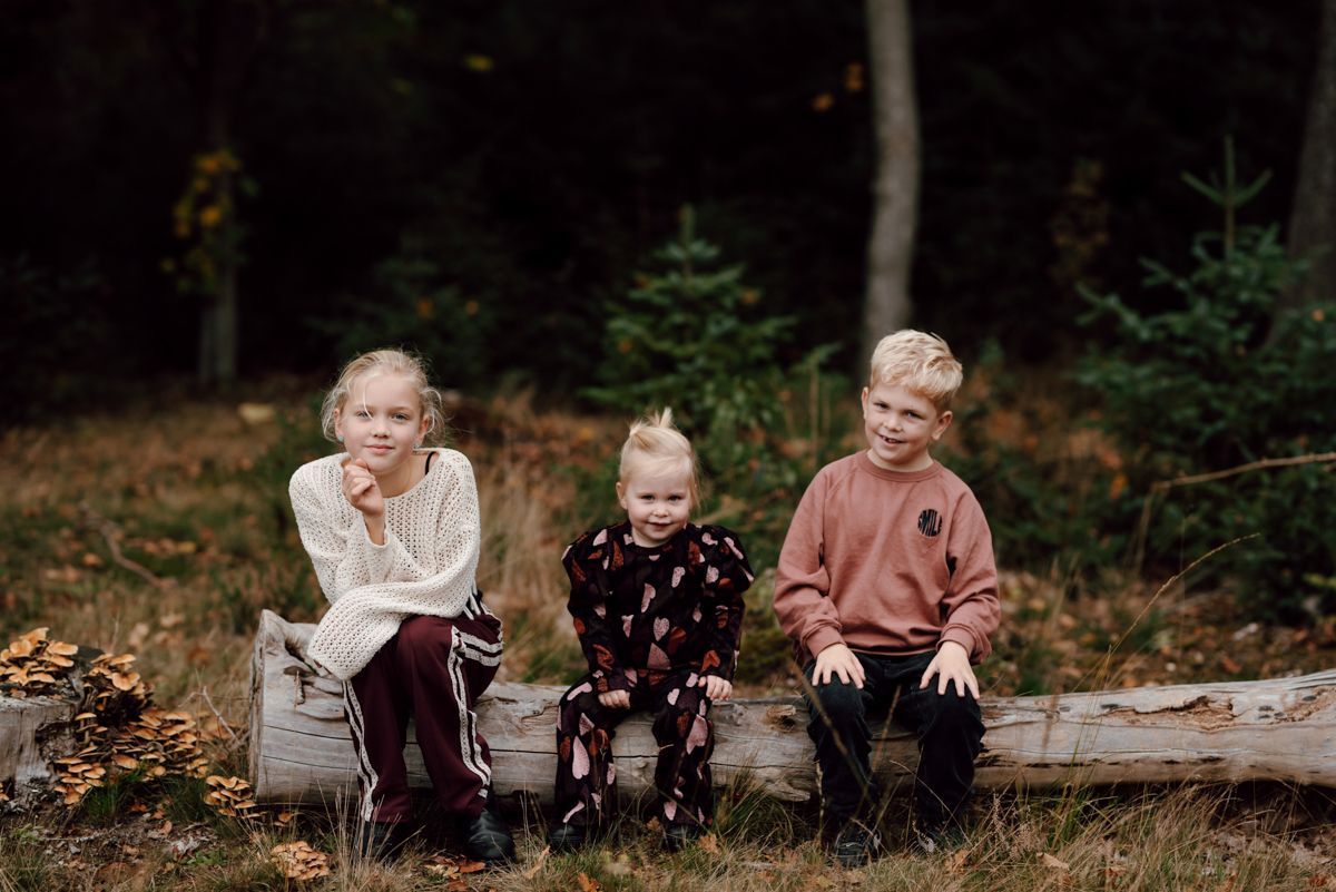 Drie kinderen zitten naast elkaar op een omgevallen boomstam in een bosrijk gebied en lachen naar de camera tijdens een fotoshoot in Appelscha