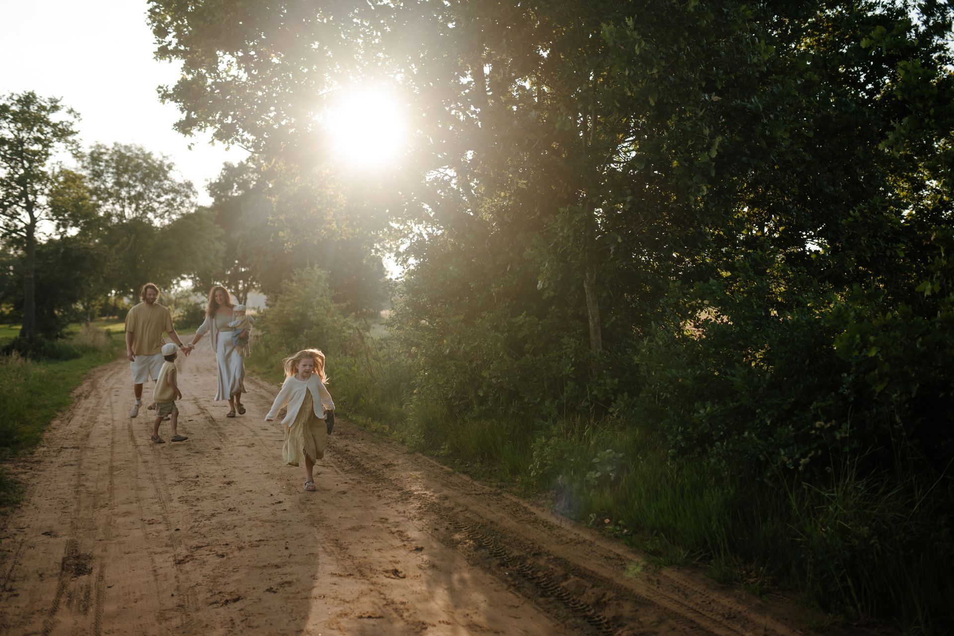 Een gezin loopt over een onverharde weg tijdens zonsondergang, waarbij het felle zonlicht door de bomen schijnt.