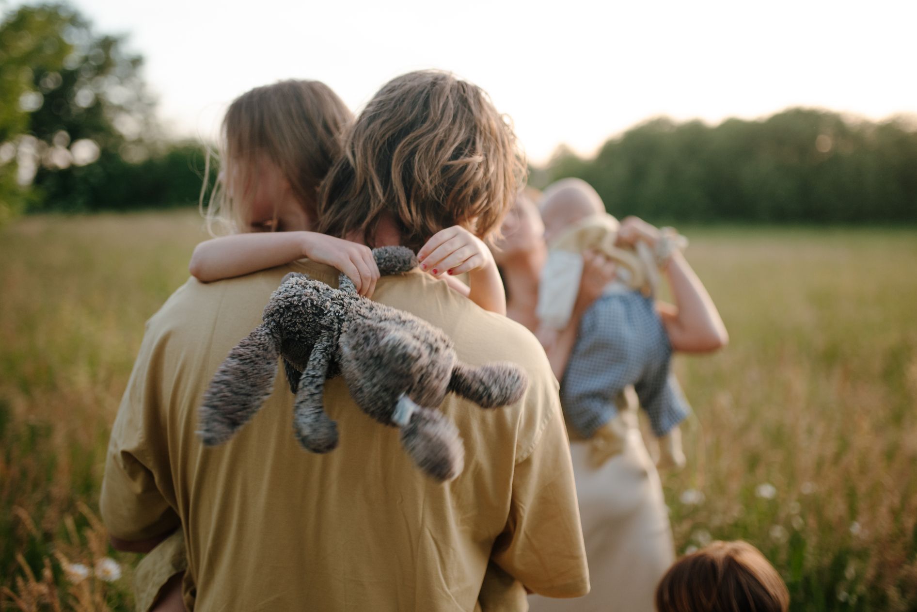 Een persoon in een geel shirt houdt een klein kind vast dat een knuffeldier vasthoudt, in een grasveld tijdens het gouden uur.