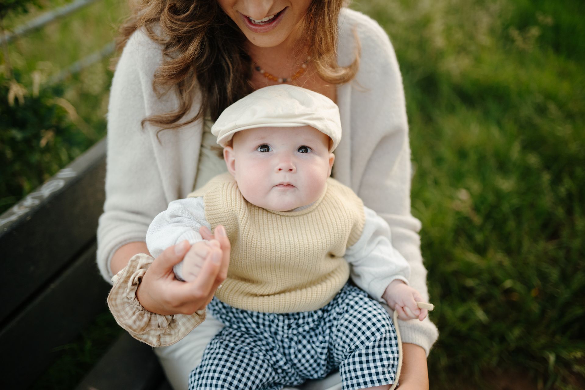 Een moeder houdt een baby vast, gekleed in een licht gebreid vestje, een broek met patroon en een platte pet, terwijl ze op een houten bankje zit.