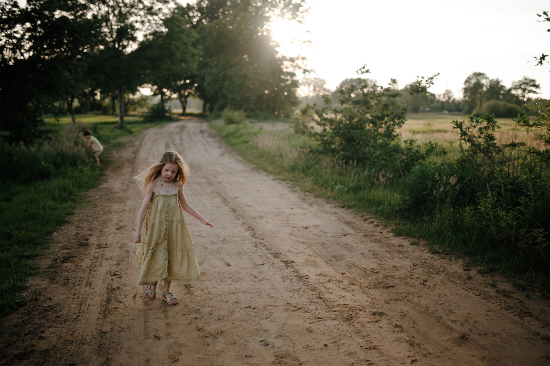 Een kind in een lichtgekleurde jurk loopt over een zandpad in een landelijke, zonnige omgeving, tijdens een gezinsreportage in de natuur