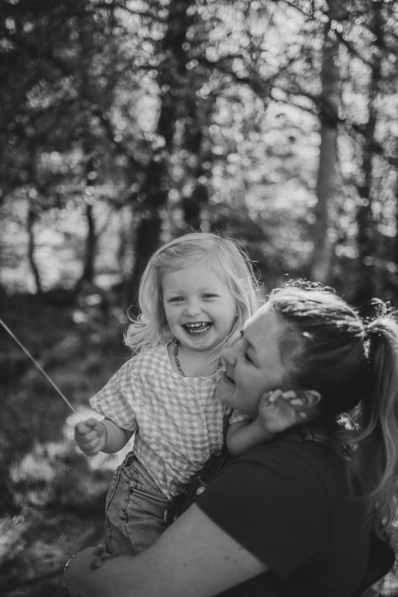 Een vrolijk kind wordt door een volwassene vastgehouden in een bosrijke omgeving, met de zon die door de bomen op de achtergrond schijnt.