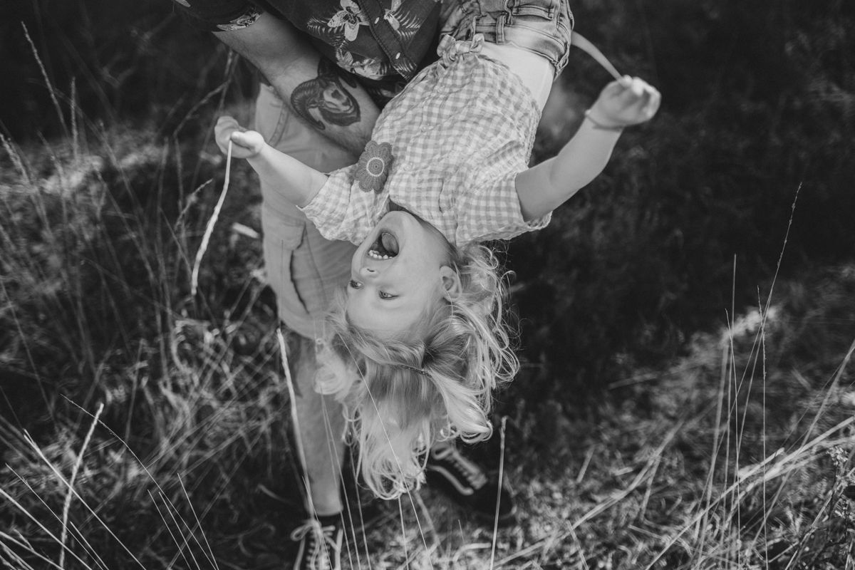 Op deze zwart-witfoto houdt iemand een lachend kind ondersteboven vast in een grasveld in de buitenlucht tijdens een gezinsshoot
