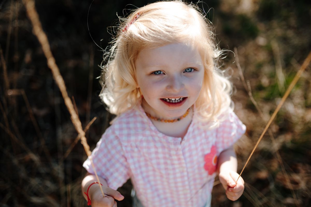 Een lachend jong kind met blond haar, gekleed in een lichtroze geruit shirt, staat buiten en houdt dunne grassprietjes vast tijdens een fotoshoot