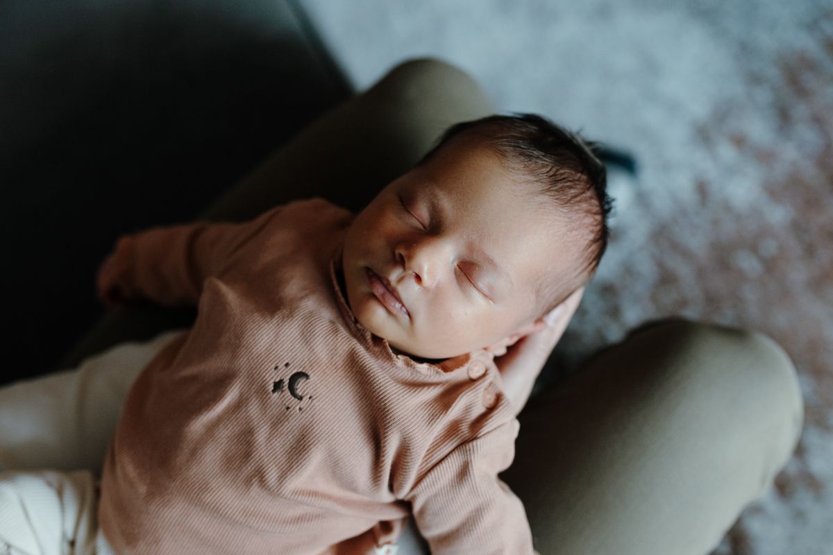 Een slapende pasgeborene in een beige rompertje ligt op iemands schoot, met op de achtergrond een zachte deken tijdens een newbornshoot
