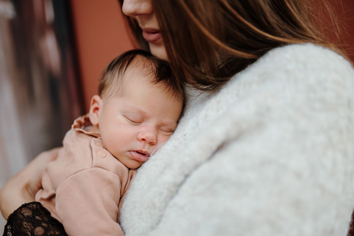 Een slapende baby wordt dicht tegen zich aan gehouden in de armen van een vrouw, gewikkeld in een zachte beige trui tijdens een newbornshoot in Groningen