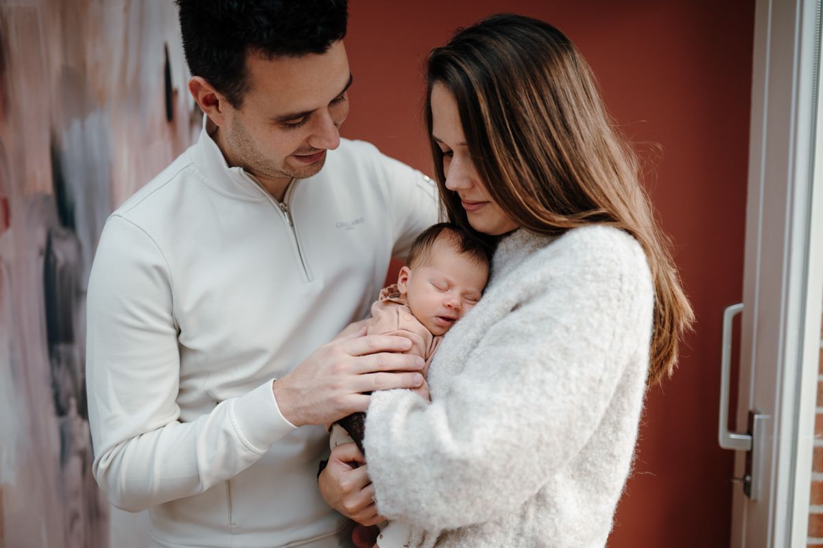 Twee volwassenen wiegen voorzichtig een slapende baby in een deuropening en kijken glimlachend naar het kind tijdens een newbornshoot in Groningen
