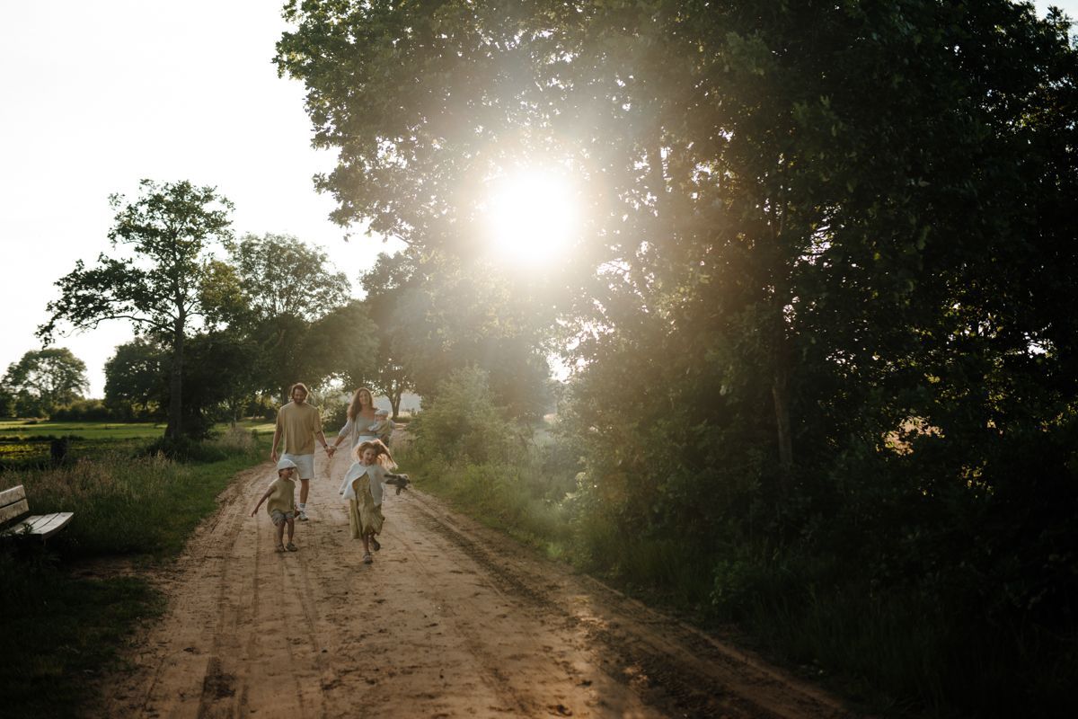 Een gezin loopt over een zandpad richting de zon, omringd door bomen op een heldere, nevelige dag.