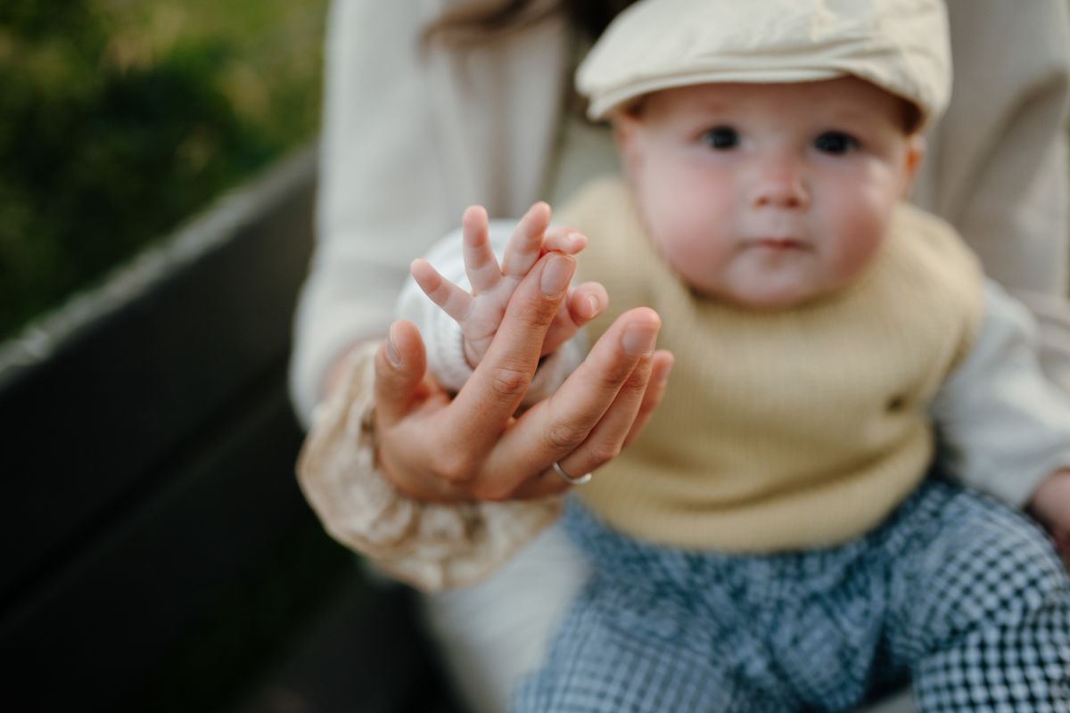 Een persoon houdt het kleine handje van een baby vast, terwijl de baby, met een mutsje en trui aan, buiten op schoot zit.
