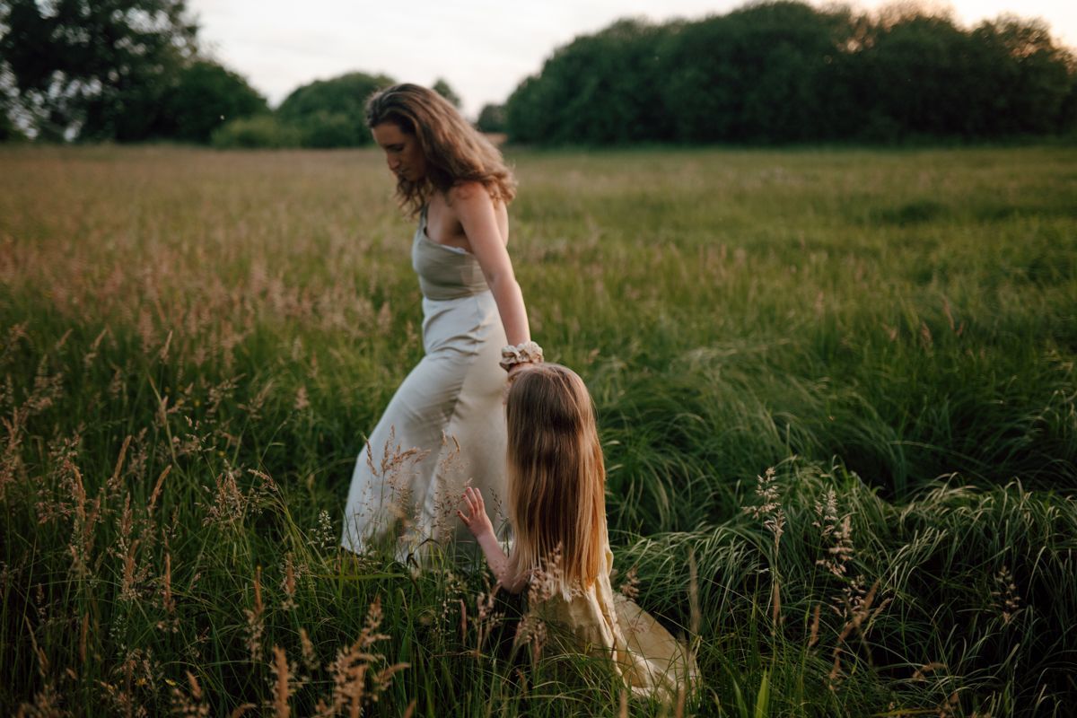 Een moeder leidt haar dochter aan de hand door een hoog grasveld in de schemering.