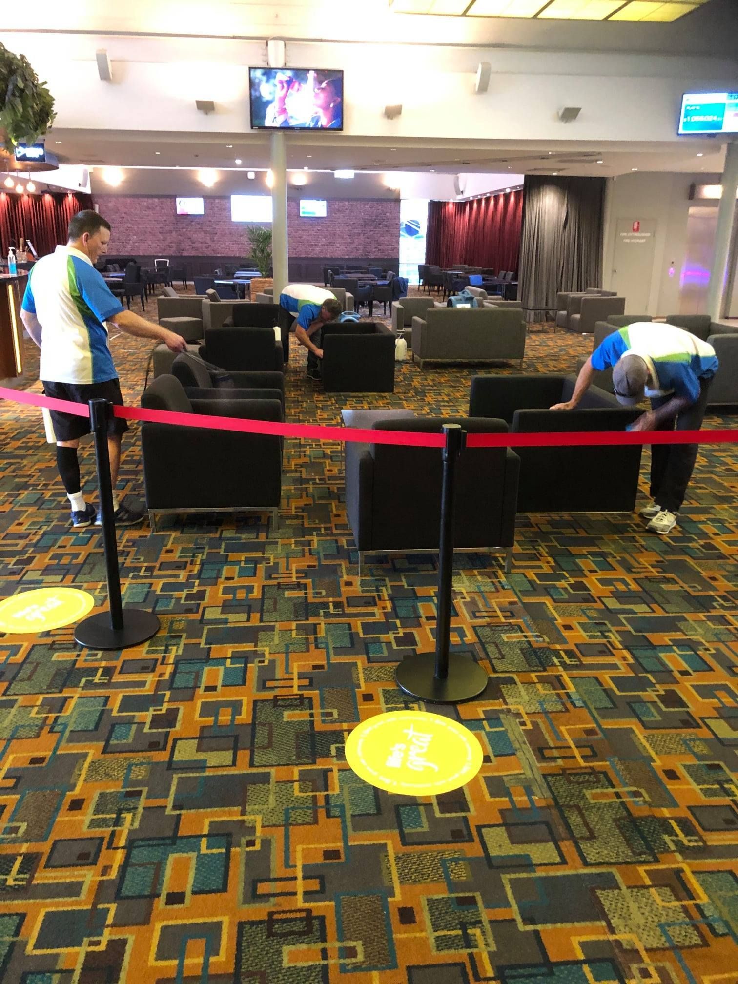 Two staff members in casual uniforms rearrange black chairs in a spacious lounge with patterned carpeting.