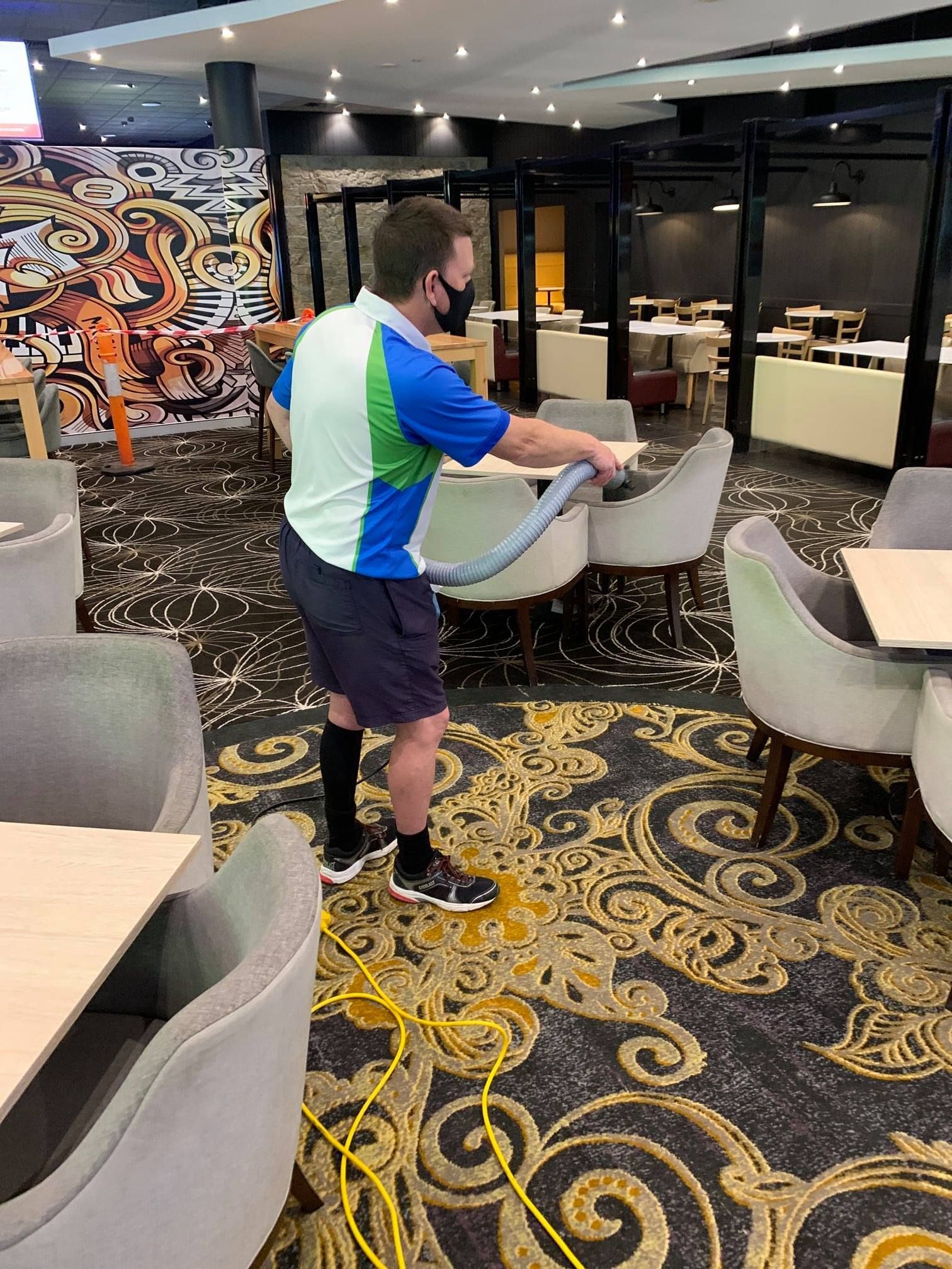 A person in a blue and green polo and shorts vacuums a patterned carpet in a restaurant dining area.